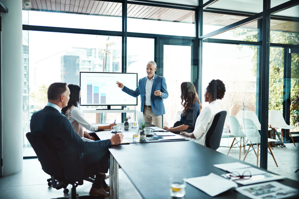 A corporate meeting for the installation of an enterprise knowledge management plan where a leader discusses the details of the plan while presenting figures in front
