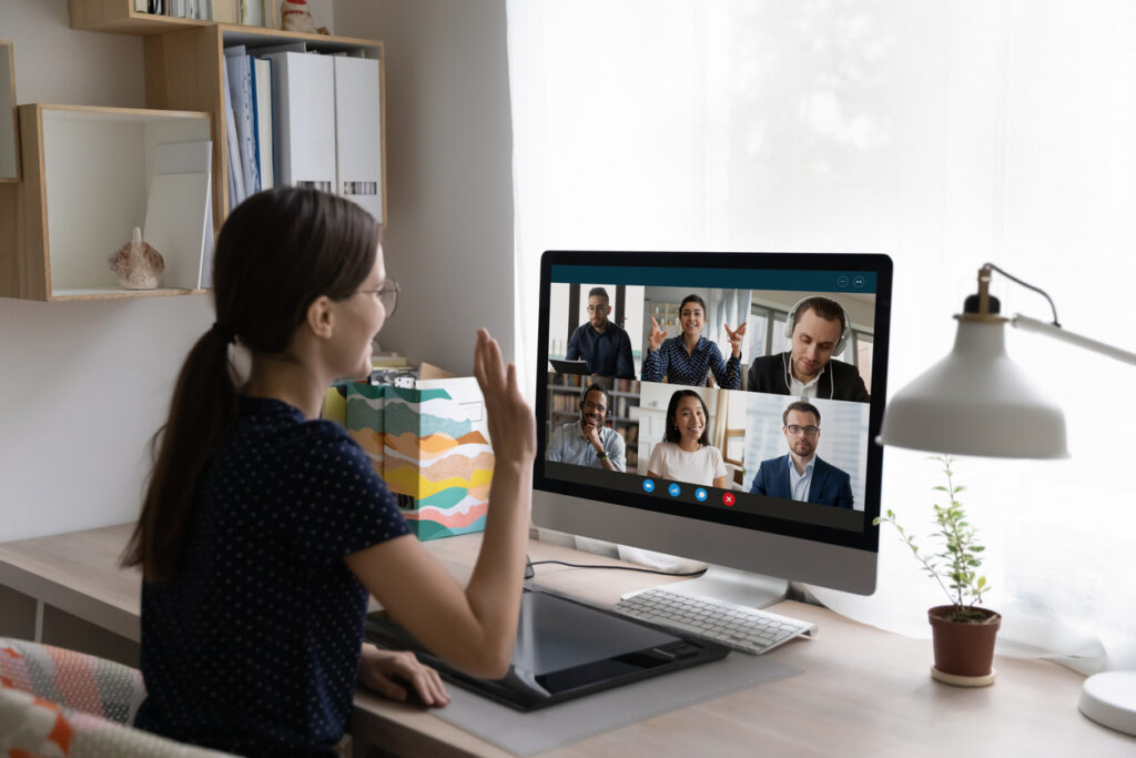 Young woman sit at desk in front of pc, looks at screen, wave hand greeting diverse, multinational colleagues during an online team building that allows for knowledge-sharing activities.