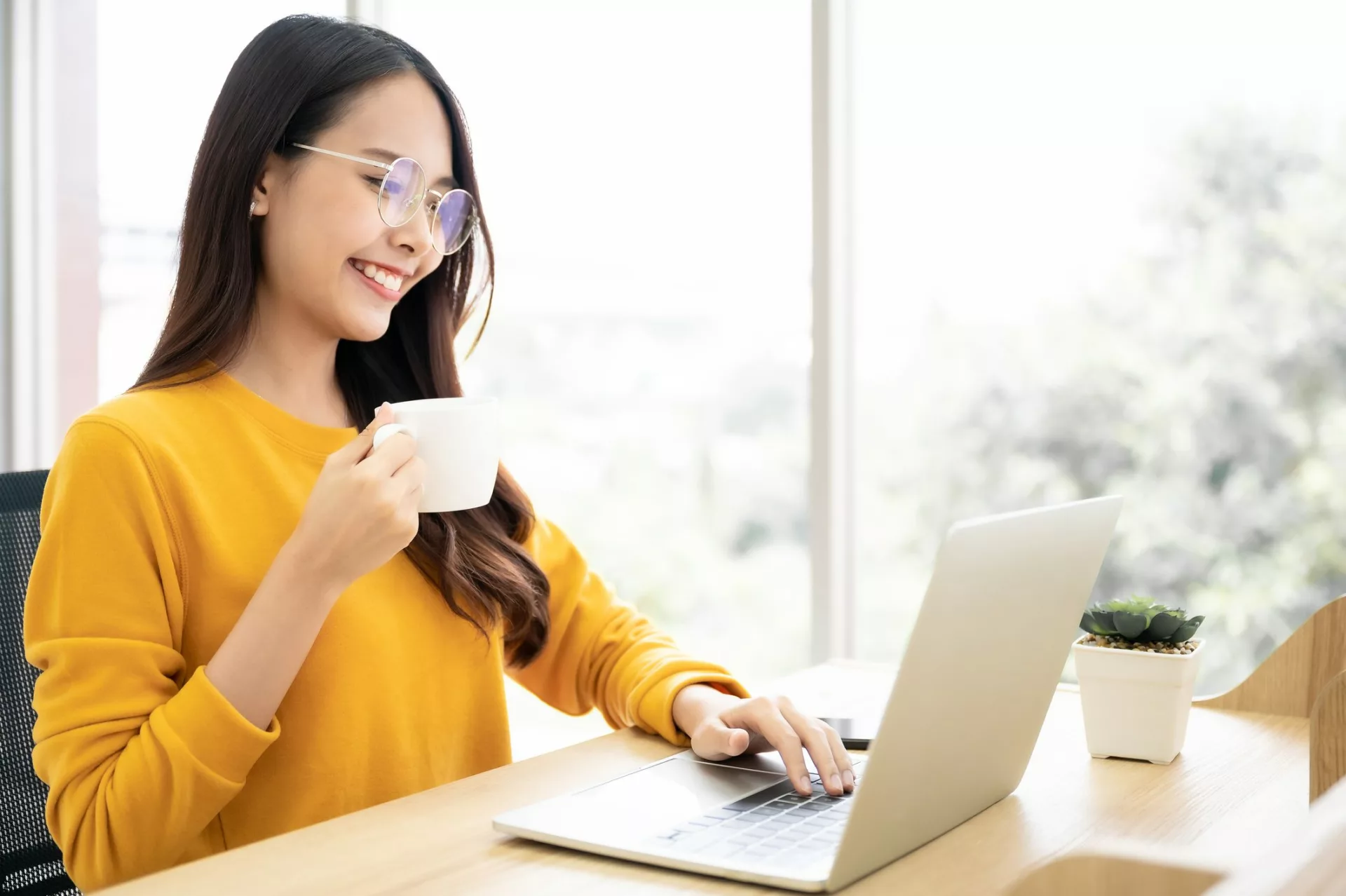 woman in yellow sweater with coffee smiles while browsing the best knowledge base software on her laptop