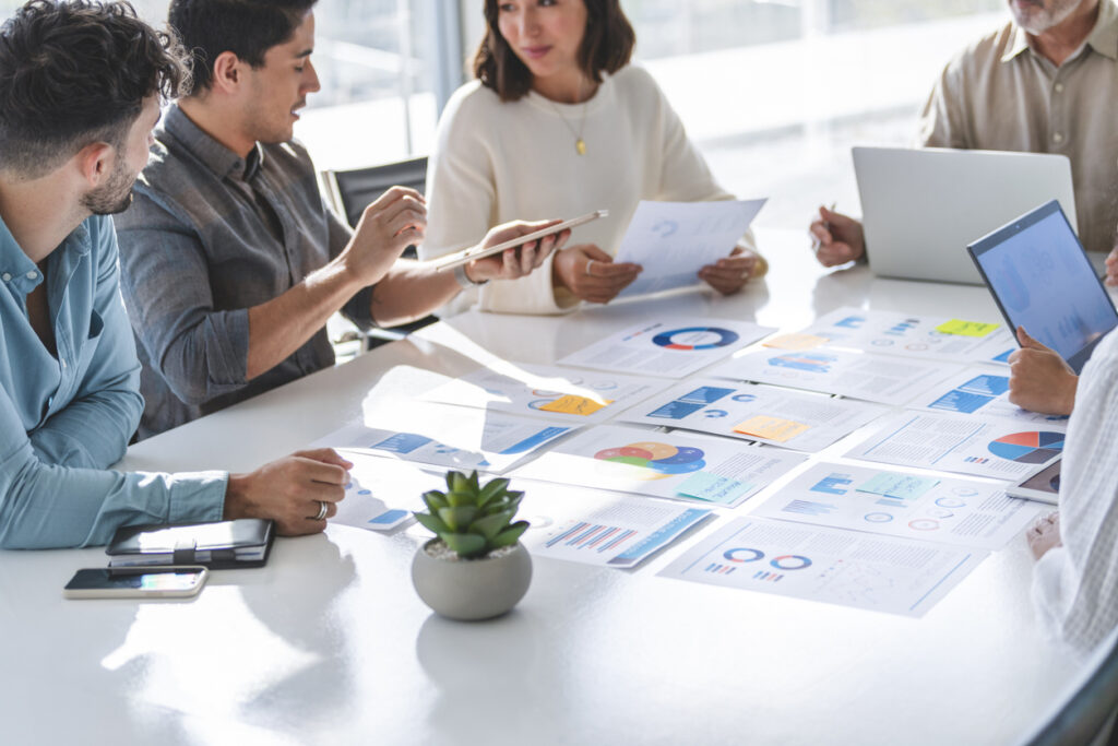 A product development team collaborates around a table filled with charts and reports, using shared insights and data to accelerate product development through Enterprise Intelligence.